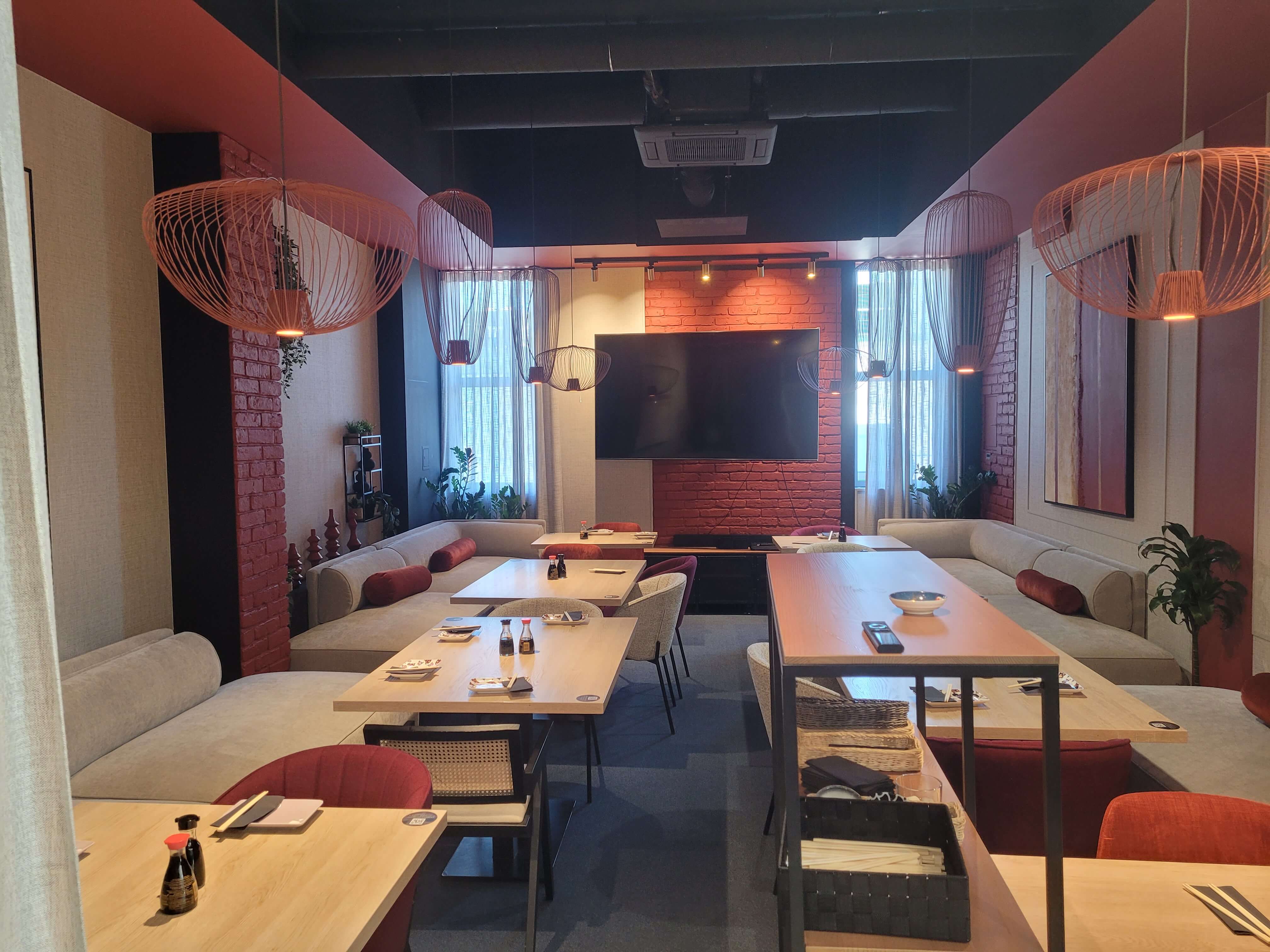 Strefa restauracji z beżowymi sofami, ceglaną ścianą, telewizorem i ażurowymi lampami wiszącymi
