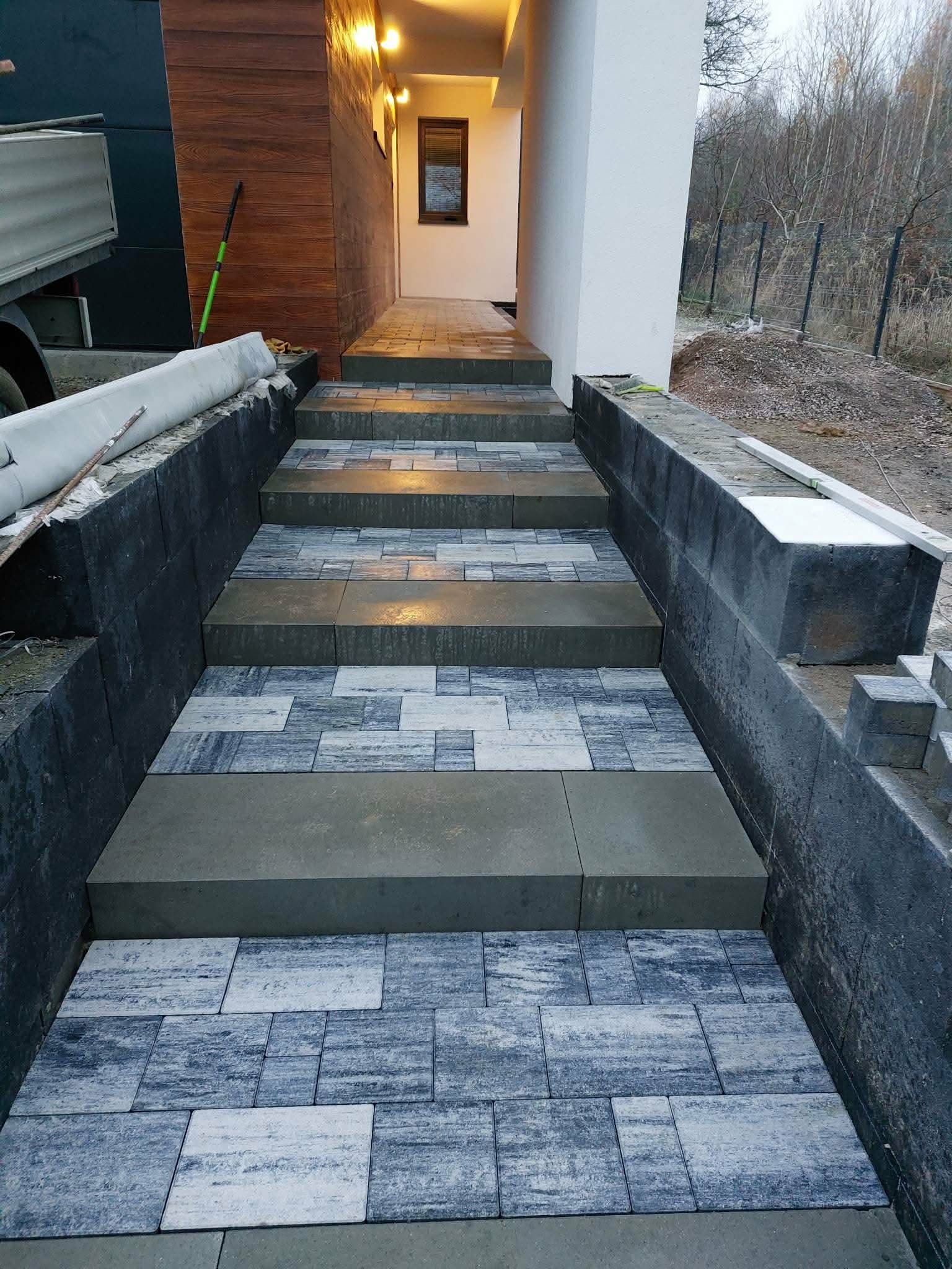 Zewnętrzne szerokie schody wykończone szaro-melanżową kostką brukową z betonowymi podstopnicami, prowadzące do wejścia domu