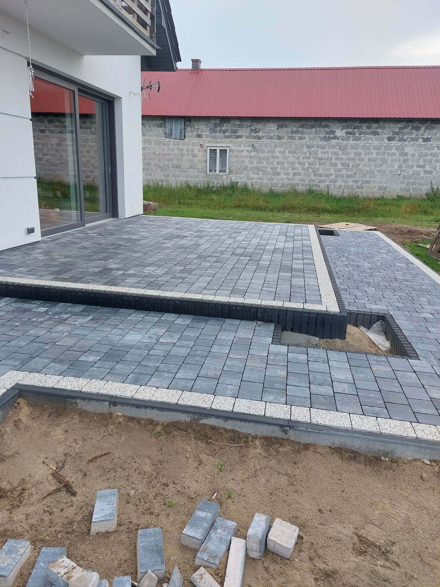 Dwupoziomowy taras z szarej kostki brukowej z jasnym obrzeżem i ciemnymi palisadami, przy domu z białą elewacją