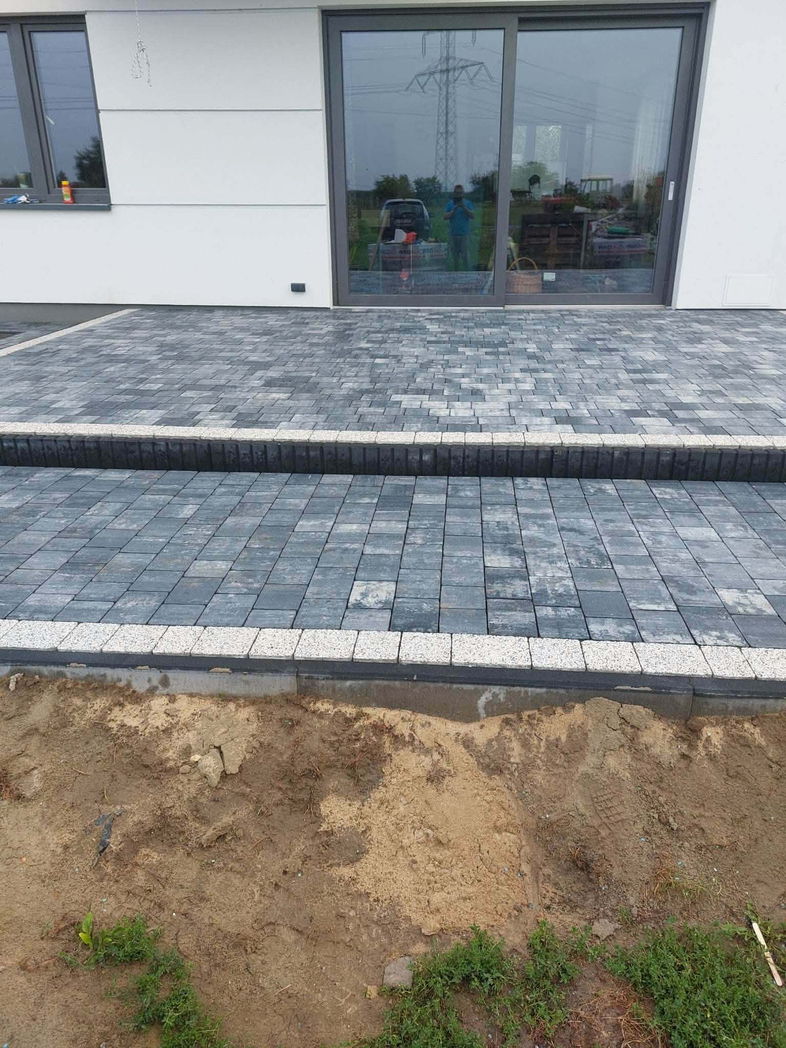 Taras z antracytowej kostki brukowej z dwoma poziomami i jasnym granitowym obrzeżem, przy przeszklonej ścianie domu